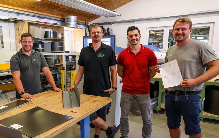 Vier Männer stehen lächelnd in einer Werkstatt, einer hält ein Blatt Papier, auf dem Tisch liegen Metallmaterialien.