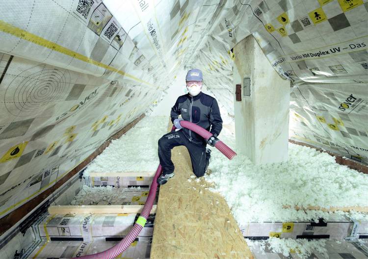 Ein Arbeiter in Schutzkleidung installiert eine Isolierung auf einem Dachboden, umgeben von weißem Isoliermaterial.