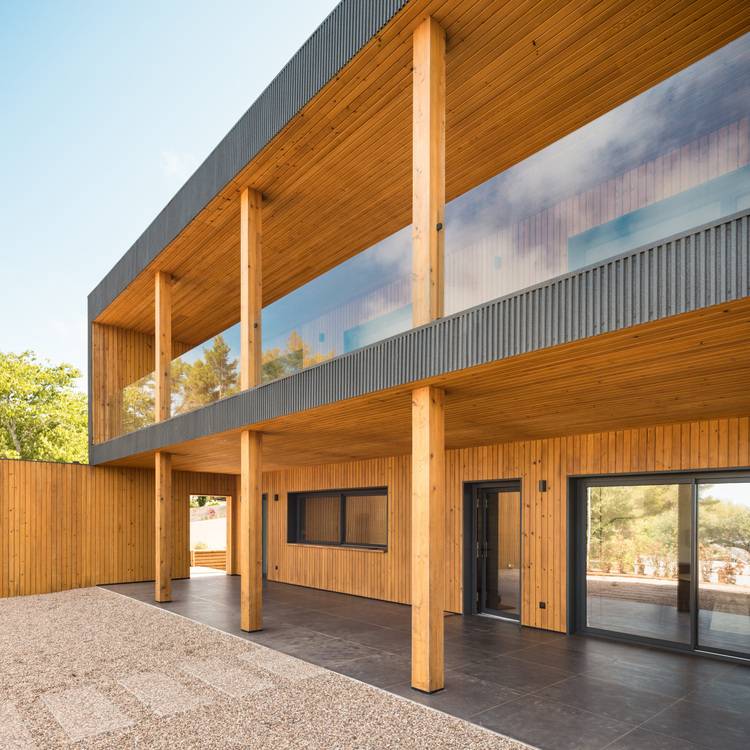 Modernes Holzhaus mit großen Fenstern und Balkon, umgeben von Bäumen und einer Kiesterrasse.