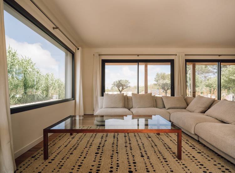 Ein geräumiges Wohnzimmer mit einem großen beigen Sofaelement, einem Couchtisch aus Glas und großen Fenstern mit Blick ins Grüne.