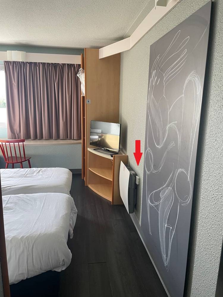 Hotelzimmer mit zwei Betten, Fernseher, abstrakter Wandkunst und einem roten Pfeil, der auf ein kleines Gerät auf dem Boden zeigt.