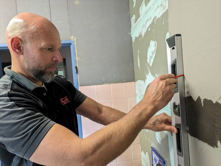 Glatzköpfiger Mann überprüft mit einer Wasserwaage die Ausrichtung einer Wand in einem teilweise gefliesten Raum.