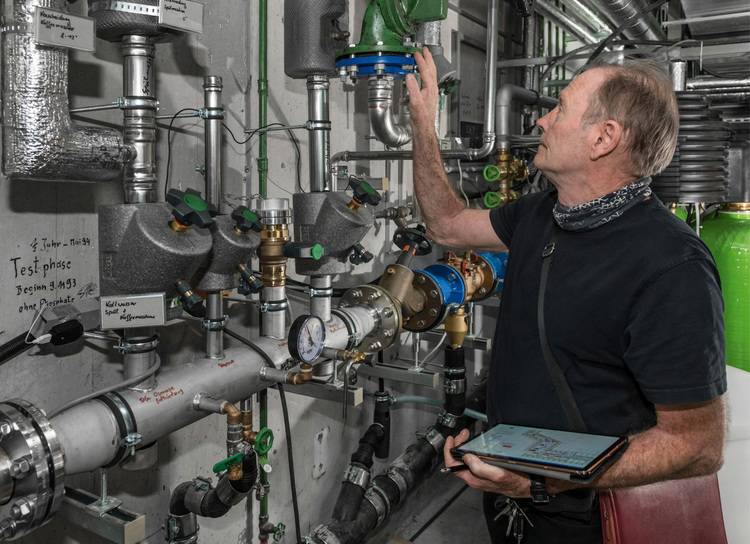 Ein SHK-Handwerker überprüft mit einem Tablet in der Hand industrielle Rohrleitungen und Ventile in einem Technikraum, um die Effizienz der Gebäudetechnik zu optimieren.