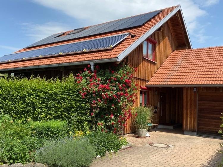 Ein Holzhaus mit Solarzellen auf dem roten Dach, umgeben von Grün und blühenden roten Blumen.