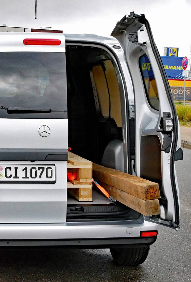 Alt-Text: „Transporter für Baumaterialien, der vor einer Baustelle parkt; die Ladeklappe ist offen und lange Holzträger sind herausgeschoben, betont wird effiziente Logistik bei Bauvorhaben in der Gebäudetechnik.“.