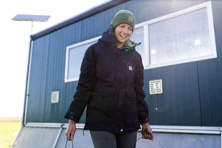 Eine Person in einer schwarzen Jacke und einer grünen Mütze steht lächelnd vor einem grünen Gebäude mit großen Fenstern.