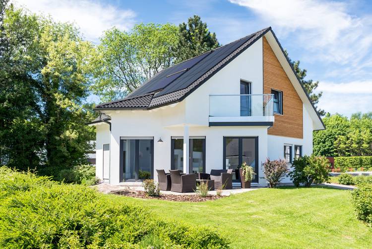 Alt-Text: „Modernes Einfamilienhaus mit Solaranlagen auf dem Dach, einer Terrasse mit Sitzmöbeln, gepflegtem Rasen und umgebenden Bäumen unter klarem Himmel, symbolisiert nachhaltige Bauweise und Energieeffizienz im Gebäudetechniksektor.