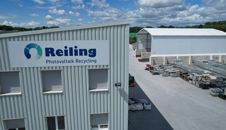 ALT-Text: „Außenansicht der Reiling Photovoltaik-Recyclinganlage mit Lagerhalle und gestapelten Materialien, umgeben von Wolkenhimmel – Fokus auf nachhaltige Energieeffizienz und Recyclinglösungen in der Gebäudetechnikbranche.