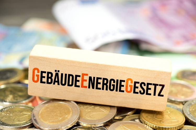 Holzblock mit der Aufschrift 'Gebäudeenergiegesetz' neben gestapelten Münzen und verschwommenen Euroscheinen im Hintergrund, symbolisiert finanzielle Aspekte und gesetzliche Anforderungen im Bereich Energieeffizienz für Entscheider in der Gebäudetechnikbranche.