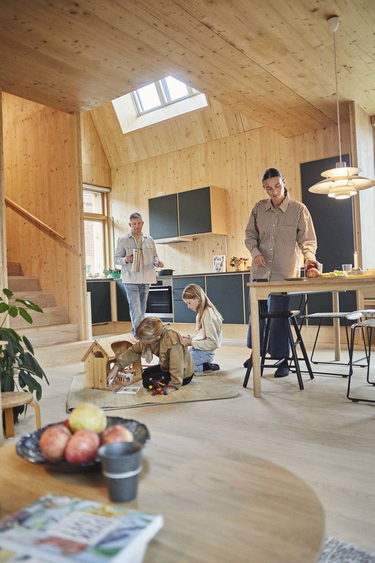 Alt-Text: „Familie nutzt energieeffiziente Holz-Küche mit moderner Heizungstechnik; zwei Kinder spielen, während zwei Erwachsene ein nachhaltiges Abendessen zubereiten.“.