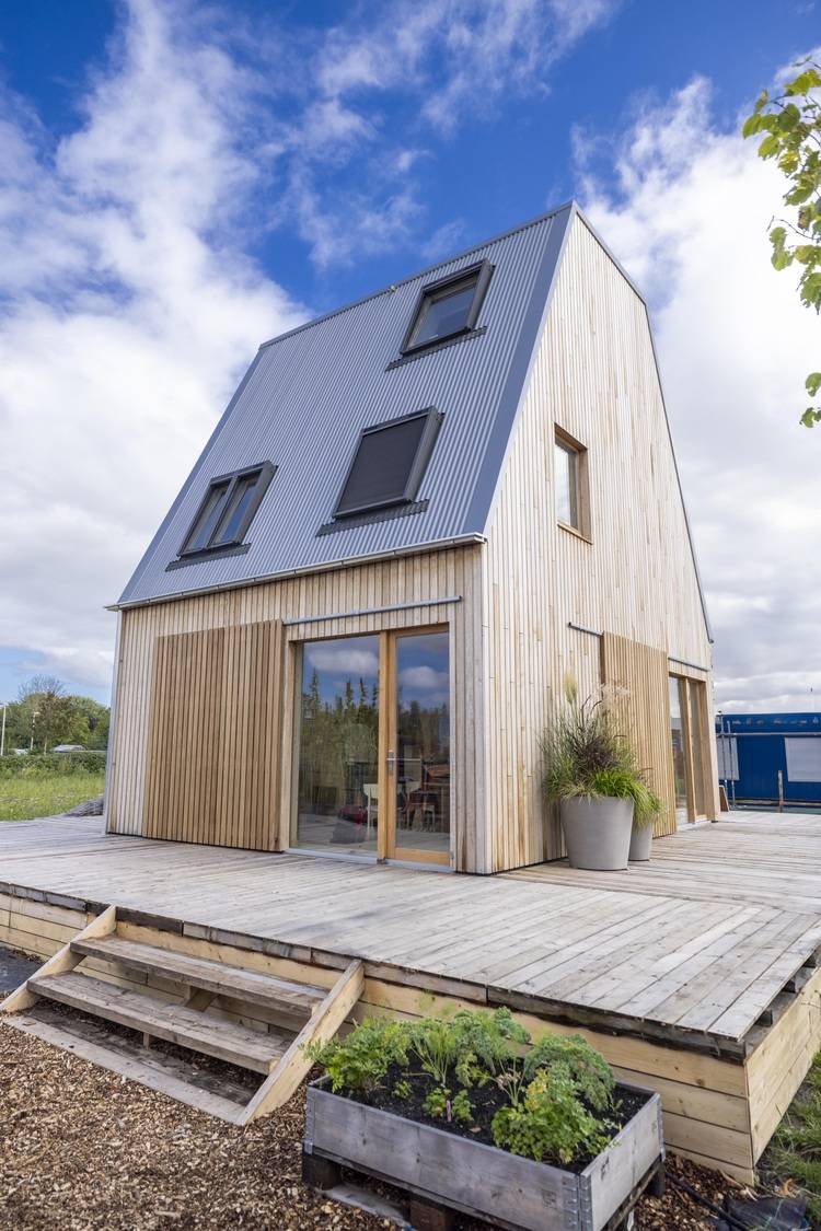 Alt-Text: „Energieeffizientes Wohnhaus mit großen Fenstern, metallischer Dacheindeckung und Holzterrasse, ideal für nachhaltige Projekte in der Gebäudetechnik.